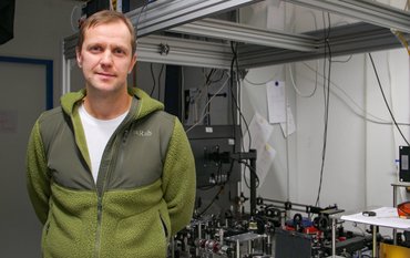Sergey Lobanov wearing a green fleece jacket stands in his laboratory next to a table on which a laser apparatus is set up
