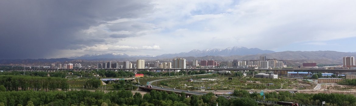 Skyline of a city in front of snowy mountains. Green trees in the foreground. From left, dark clouds arrive.