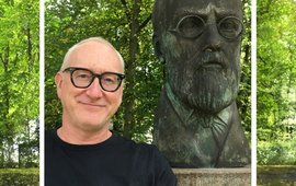 Portrait photo of a man with dark glasses next to the larger-than-life bronze bust of a man with a full beard and glasses. In the background: green trees.