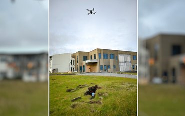 Eine Drohne fliegt in der Luft und hält ein Messystem an einem Seil über den Boden.