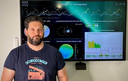 Portrait of a man in a T-shirt in front of a large screen with information on space weather.