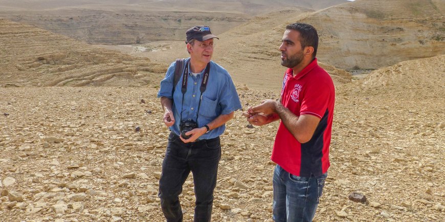 Achim Brauer and a young man talk against the backdrop of a stony and arid landscape.