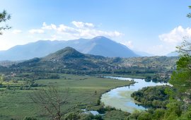 Eine grüne Landschaft. Mitte und Hintergrund: Berge. Im Vordergrund mäandert ein Fluss.