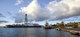 Das Forschungsschiff liegt im Hafen. Der Himmel ist bewölkt, aber blau. Das Meer ist ruhig. An Land ein asphaltierter Parkplatz mit Palmen direkt an der Hafenkante. Menschen sitzen und spazieren dort.