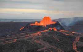 Dunkelgraue, flache Vulkanlandschaft, in der Mitte sprudelt rot-glühende Lava aus einem Krater und rinnt die Hänge entlang.