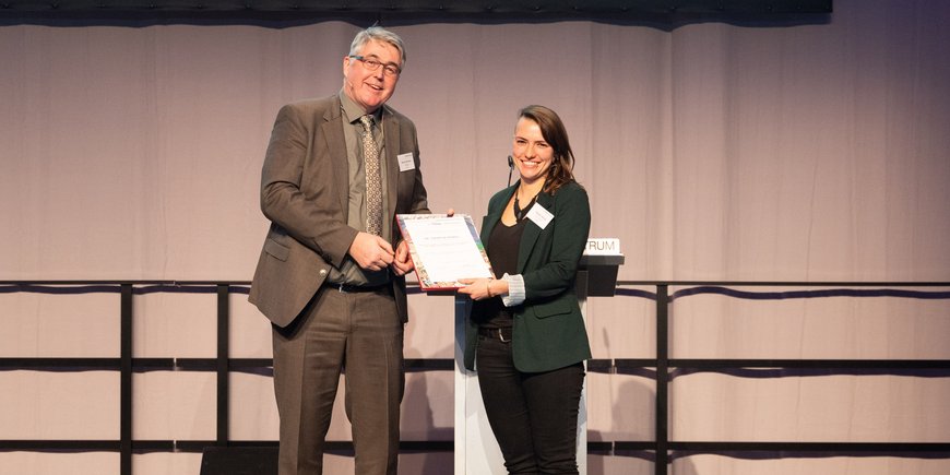 A middle-aged man wearing a brown suit hands a certificate to a young woman wearing a green jacket and black trousers. Both are standing on a black stage. A grey curtain in the background.