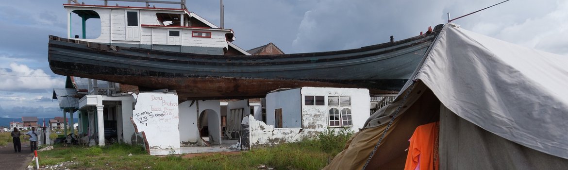 A boat is on the roof of a destroyed house, a tent on the ground, a puddle, clothes hanging on a line, two men standing next to it.