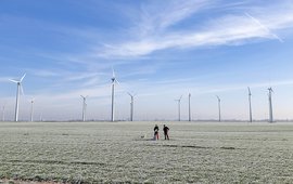A few wind turbines on flat land. Two people are standing in front of them on the frosted field. Blue sky with hazy clouds.