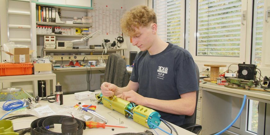 A young man sits at a laboratory table with cables, other material and diverse tools. He screws on a tube-shaped apparatus.