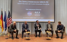 Panel discussion of five people in front of a screen. On the side a German, European and American flag each