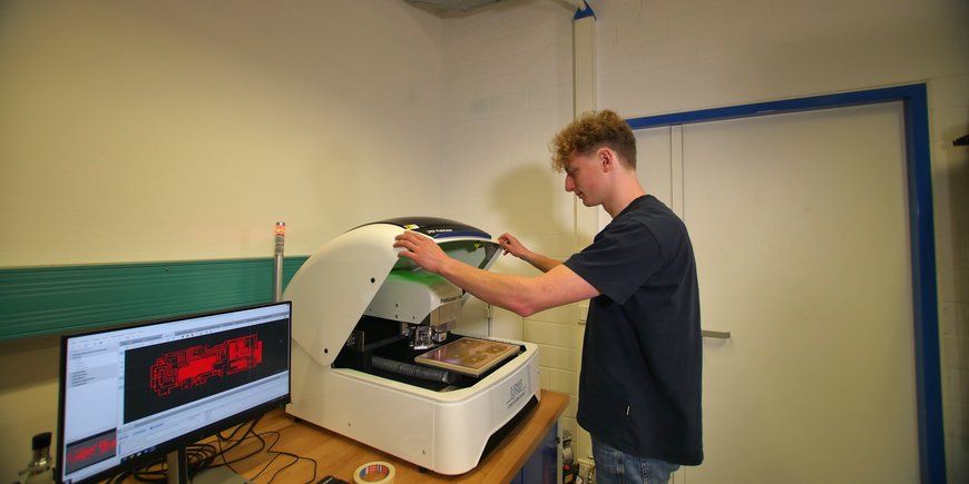 A young man stands in a laboratory at a machine that produces circuit boards. Next to it is a computer screen showing the design of the circuit board.