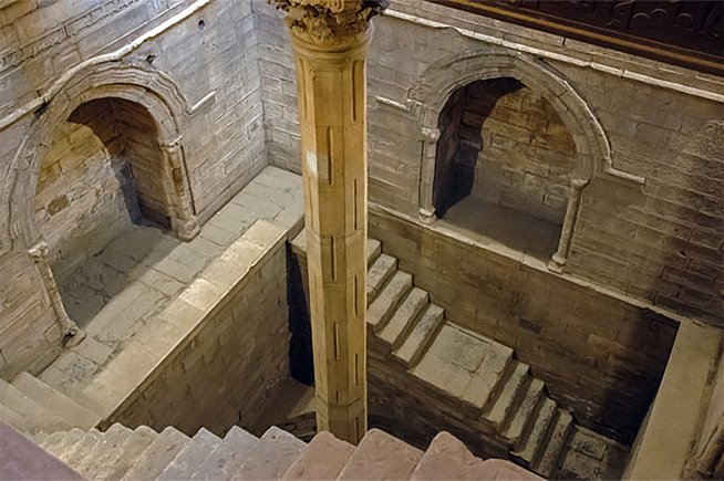 Rechteckiger gemauerter Tiefbrunnen mit einer Säule in der Mitte und Stufen, die in die Tiefe führen.