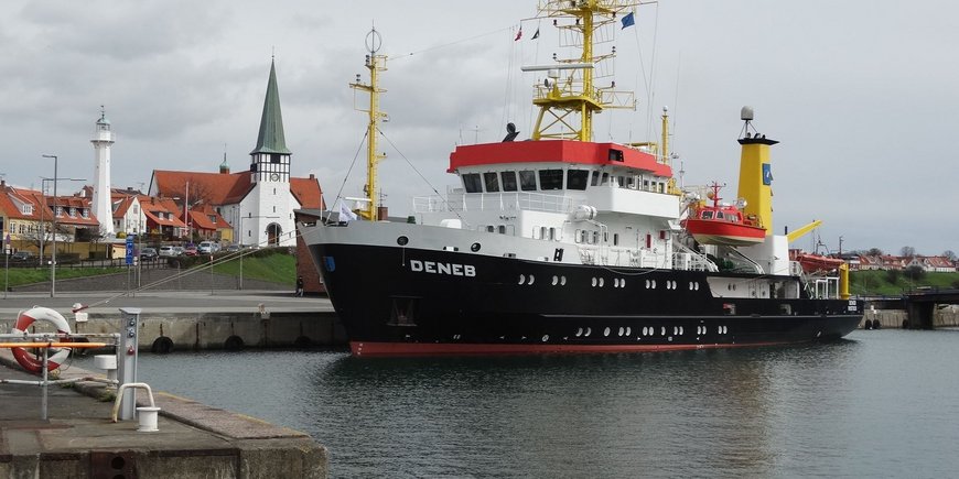 This photo shows the German survey ship "Deneb" in the harbor of the Danish town of Bornholm. The viewer looks diagonally from the front at the bow and port side of the ship from the harbor basin. The ship is about 100 meters away from the viewer. It is moored along its starboard side to the harbor wall and you can see the church of Bornholm and houses next to it about 1 kilometer away in the background. There is a lighthouse between the houses. The ship is about 52 meters long, 11 meters wide and more than 20 meters high with the mast. It has the inscription "DENEB" on the bow.