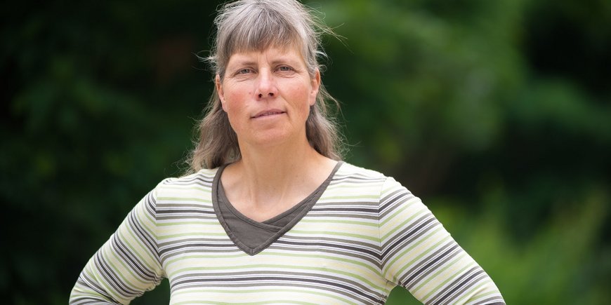 Portrait photo: A woman with long hair and fringes and a striped top stands in front of green trees.