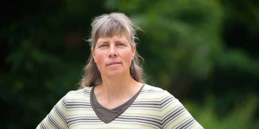 Portraitfoto: Eine Frau mit grau-blonden langen Haaren und Pony und gestreiftem Oberteil steht vor grünen Bäumen.