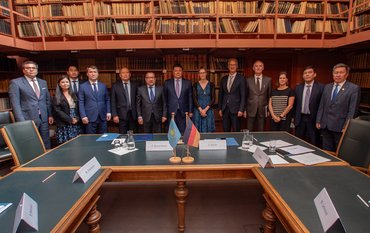 Gruppenfoto: 13 Personen stehen vor einer Bücherwand, im Vordergrund sind Tische und die Fähnchen von Kasachstan und Deutschland..