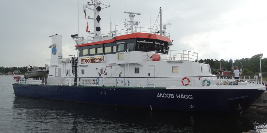 This photo shows the Swedish survey ship "Jacob Hägg" in a harbor near Gävle. It is about 36 meters long and 7.5 meters wide and 15 meters high with the mast. The ship is about 50 meters away from the viewer. The viewer looks diagonally from the edge of the harbor basin at the bow and starboard side of the ship. The ship is moored along its port side to the harbor wall. On the bow and on the side of the bridge it bears the inscription "JACOB HÄGG". The coat of arms of the Swedish Maritime Administration with an anchor and royal crown is attached to the chimney. In the background you can see the forests of the coastal region.