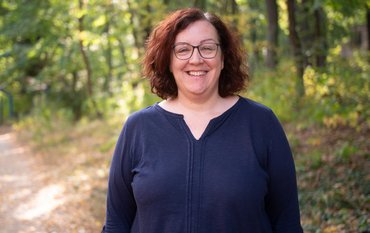 Portrait of a woman with dark, half-length hair and glasses. She is smiling and wearing a dark blue top. In the background: green forest.