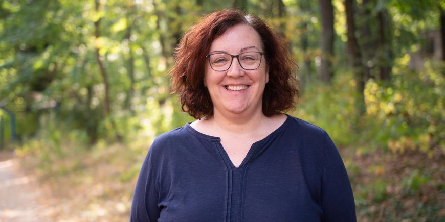 Portrait of a woman with dark, half-length hair and glasses. She is smiling and wearing a dark blue top. In the background: green forest.