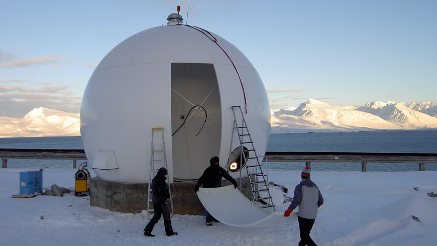 The photo shows three persons during installation works at the second antenna of the satellite receiving station Ny-Ålesund in 2005. Two persons carry the last segment of the radome, which is almost completed. Snow-covered mountains, shining brightly in the sun, are visible in the background on the opposite side of the fjord.