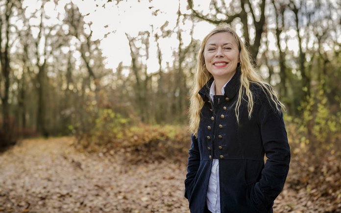 [Translate to English Portrait of a woman with long blonde hair and a blue jacket in front of treesPortrait einer Frau mit blonden langen Haaren und einer blauen Jacke vor Bäumen