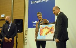 A woman and a man stand on a stage holding a picture with a coloured map of Türkiye..