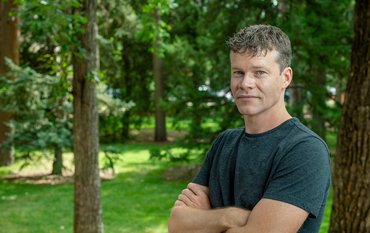 Portrait of a young man with dark hair and a black t-shirt in front of green trees.