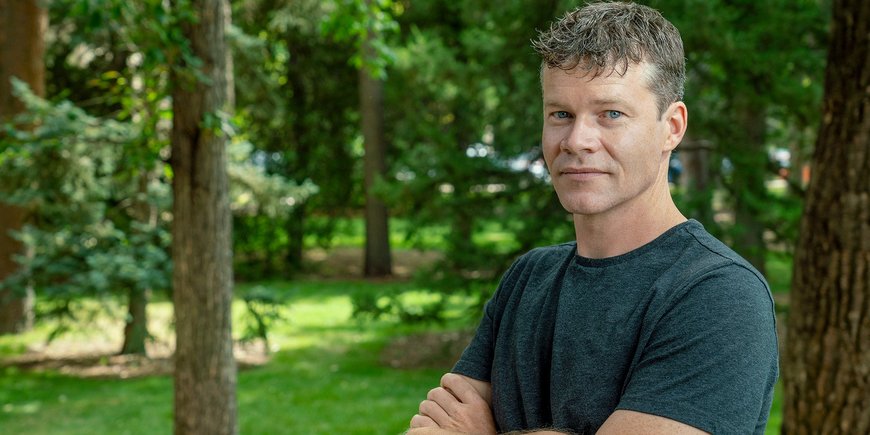 Portrait of a young man with dark hair and a black t-shirt in front of green trees.