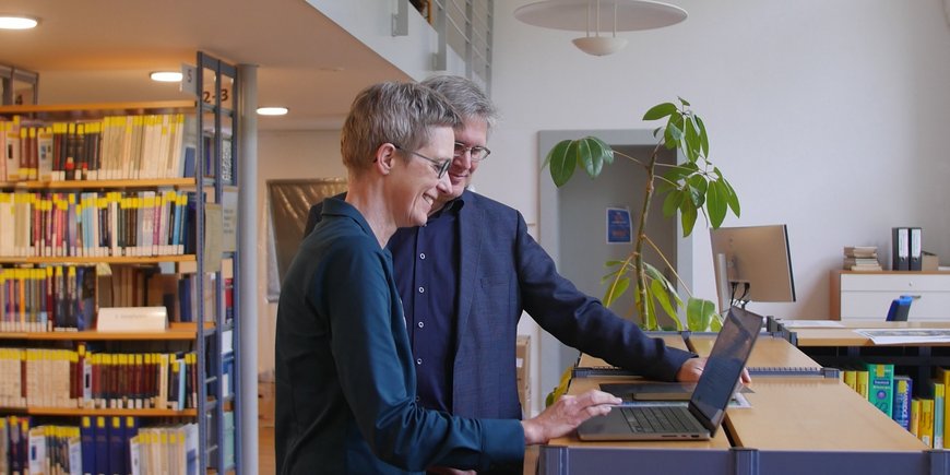 Beide Wissenschaftler:innen im Gespräch, stehend , vor einem Computer in der Bibliothek