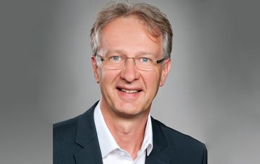 Dr. Oliver Bens, Mann mit graubraunen Haaren, hellblauen Augen und Brille, trägt einen Anzug