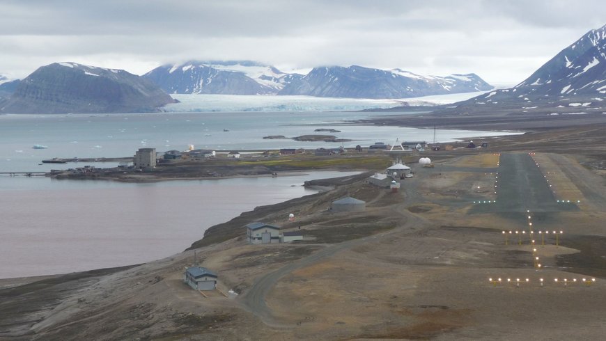 The photo shows Ny-Ålesund with the Kongsfjord, a glacier in the distance and the landing airstrip next to the receiving station.
