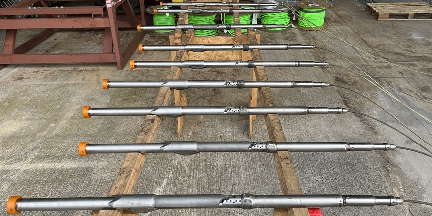 11 dünne metallische Rohre, die Bohrlochseismometer, liegen aufgereiht auf einem Holtzgestell über einem Betonboden. Im Hintergrund Kabel.