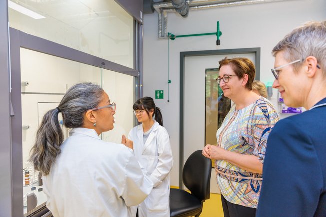 Zwei Frauen in weißem Kittel stehen an einem Laborabzug in einem chemischen Labor. Eine Frau spricht. Andere Menschen sehe und hören interessiert zu.