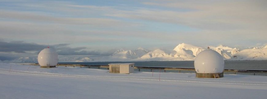 The photo shows the satellite receiving station Ny-Ålesund (NYA) with the two antennas under white radomes on the left and on the right and a cabin for receiving equipment in between.