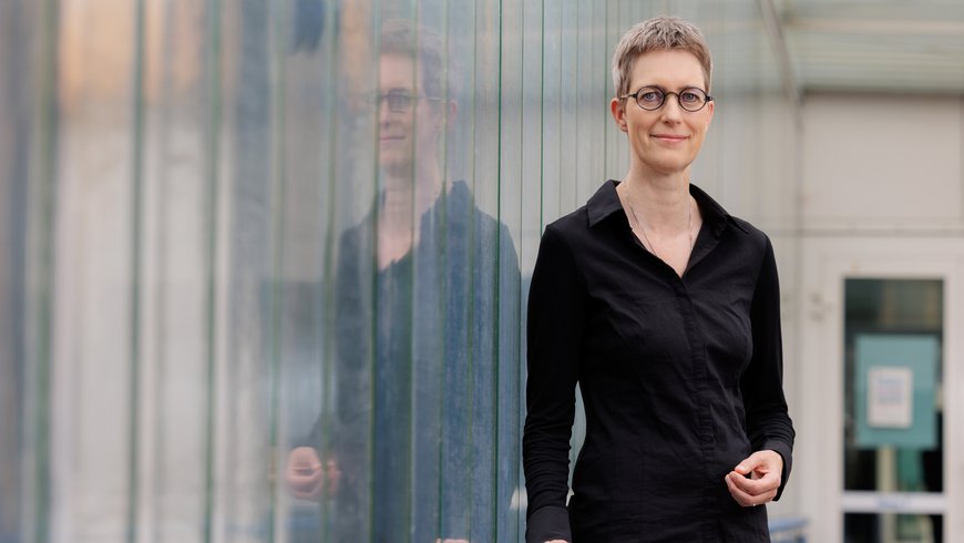 Prof. Susanne Buiter stands in a black suit in a glass passageway. The glass reflects her image.